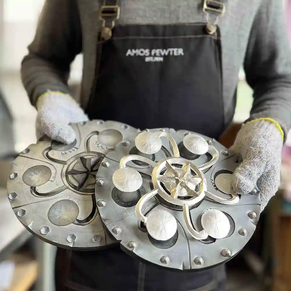 Person with an Amos Pewter apron wearing gloves holds a mold of newly poured pewter sand dollars.