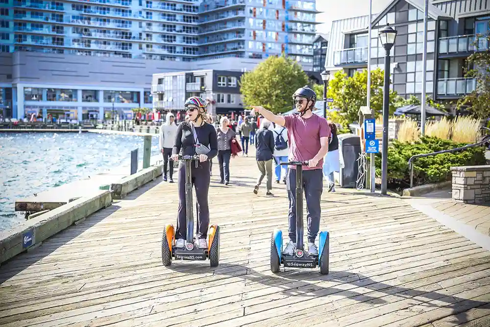 A man and a woman ride along the Halifax Waterfront aboard Segways. The man arbour points towards the harbour, the woman looks in awe.