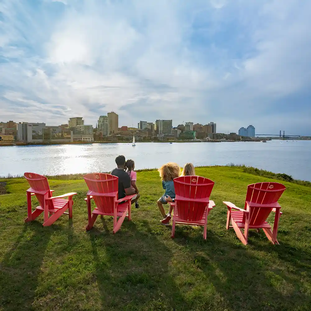 A family sit on the Parks Canada Red Chairs on Georges Island as they look towards the Halifax Skyline.