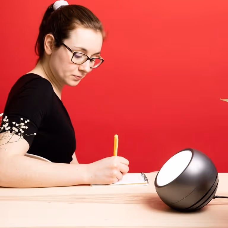 Circadian optics Leo light therapy lamp placed on a table with a person writing in a notebook next to it infront of a red wall