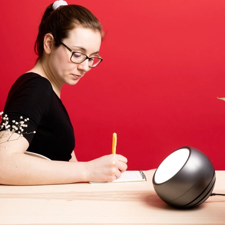 Circadian optics Leo light therapy lamp placed on a table with a person writing in a notebook next to it infront of a red wall