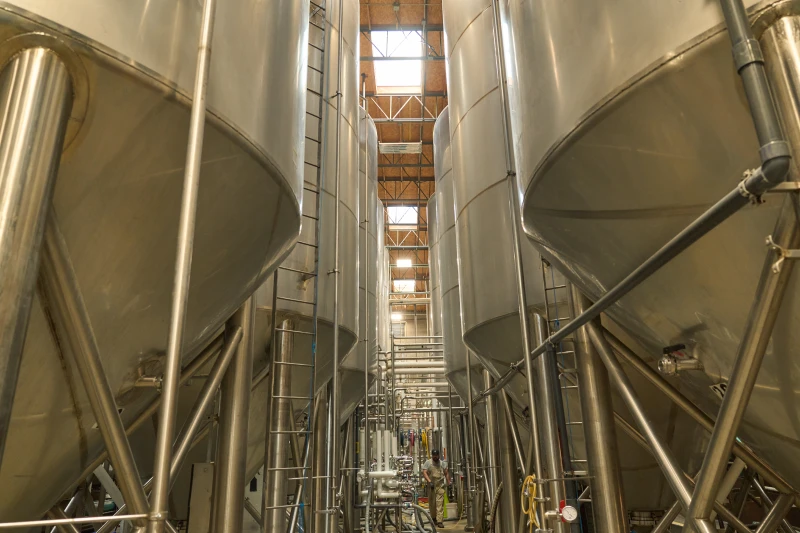 Interior view of a brewery with large stainless steel fermentation tanks and a worker among the pipes.