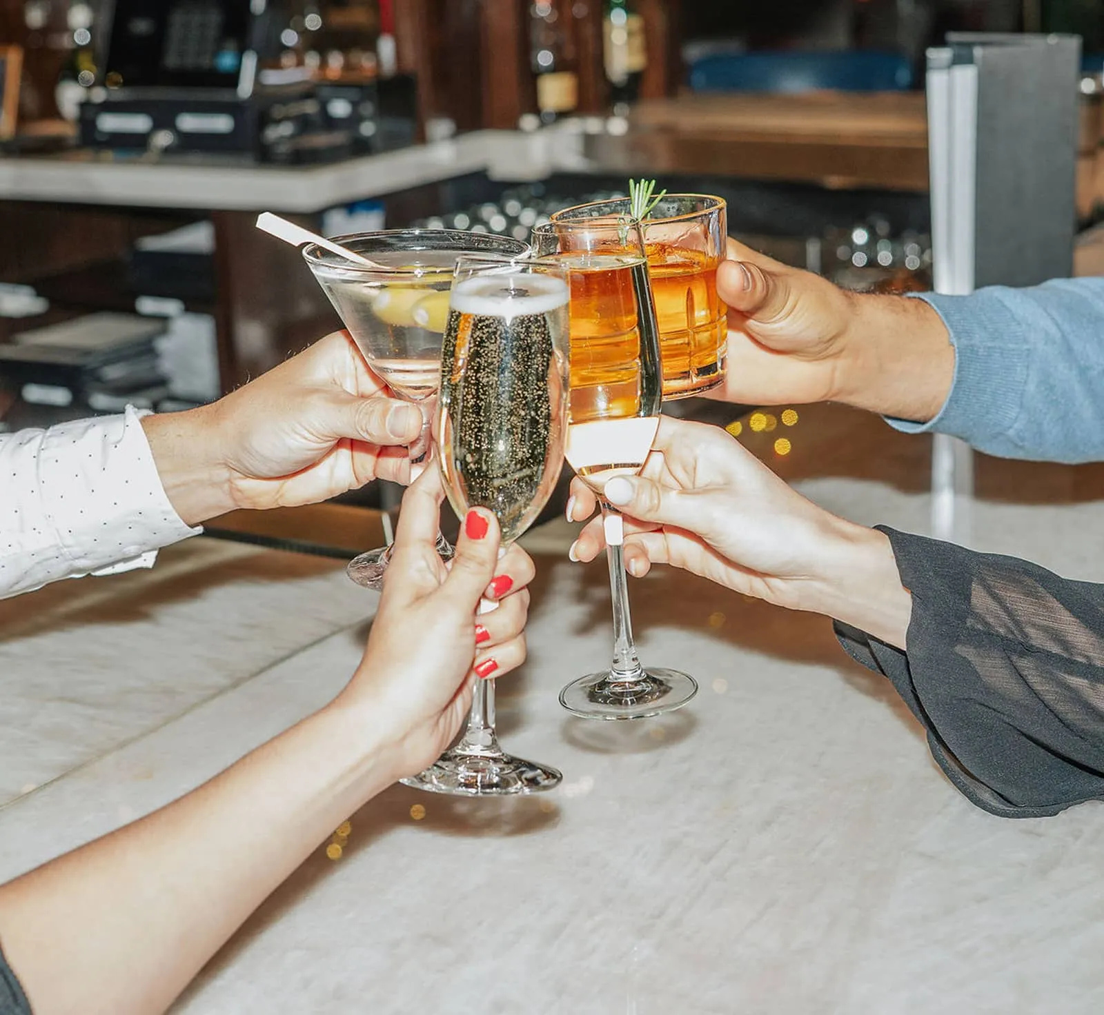 Four people clinking glasses of various cocktails and champagne at a bar counter.