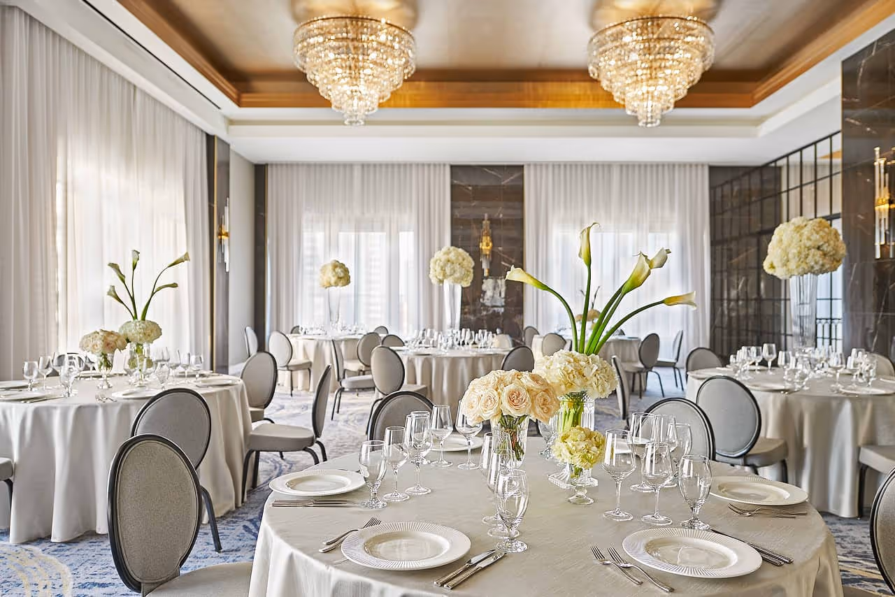 Elegant ballroom with round tables draped in beige linens, set with fine china, glassware, and floral centerpieces under crystal chandeliers.