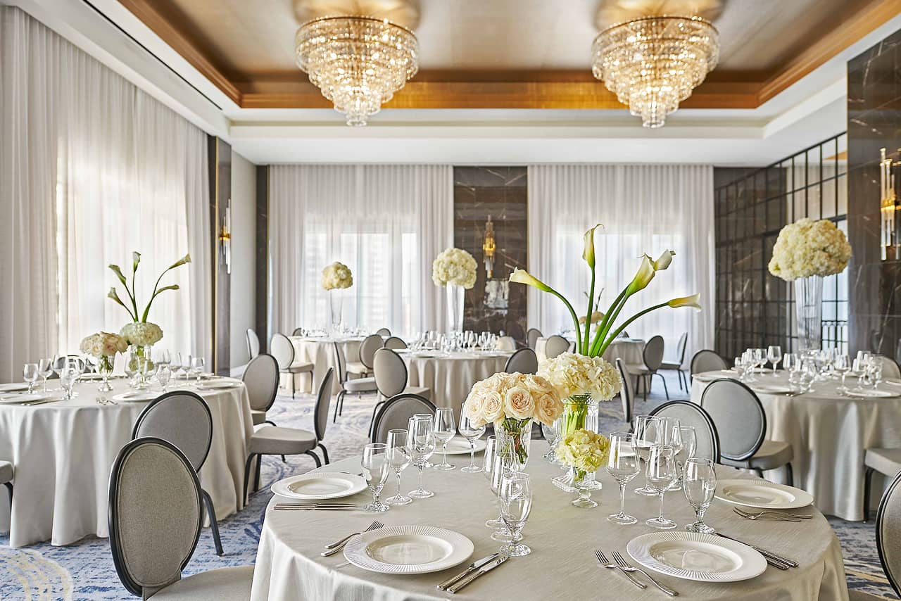 Elegant ballroom with round tables draped in beige linens, set with fine china, glassware, and floral centerpieces under crystal chandeliers.