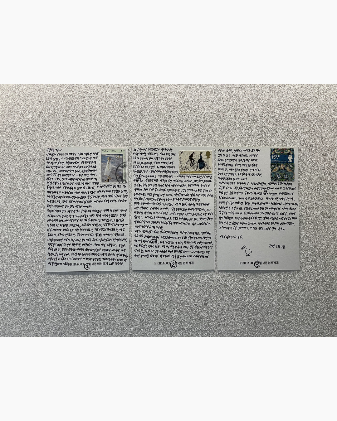 세 장의 손글씨가 쓰여진 종이 카드가 벽에 나란히 붙어 있으며, 각각 다른 우표가 상단에 부착되어 있다.