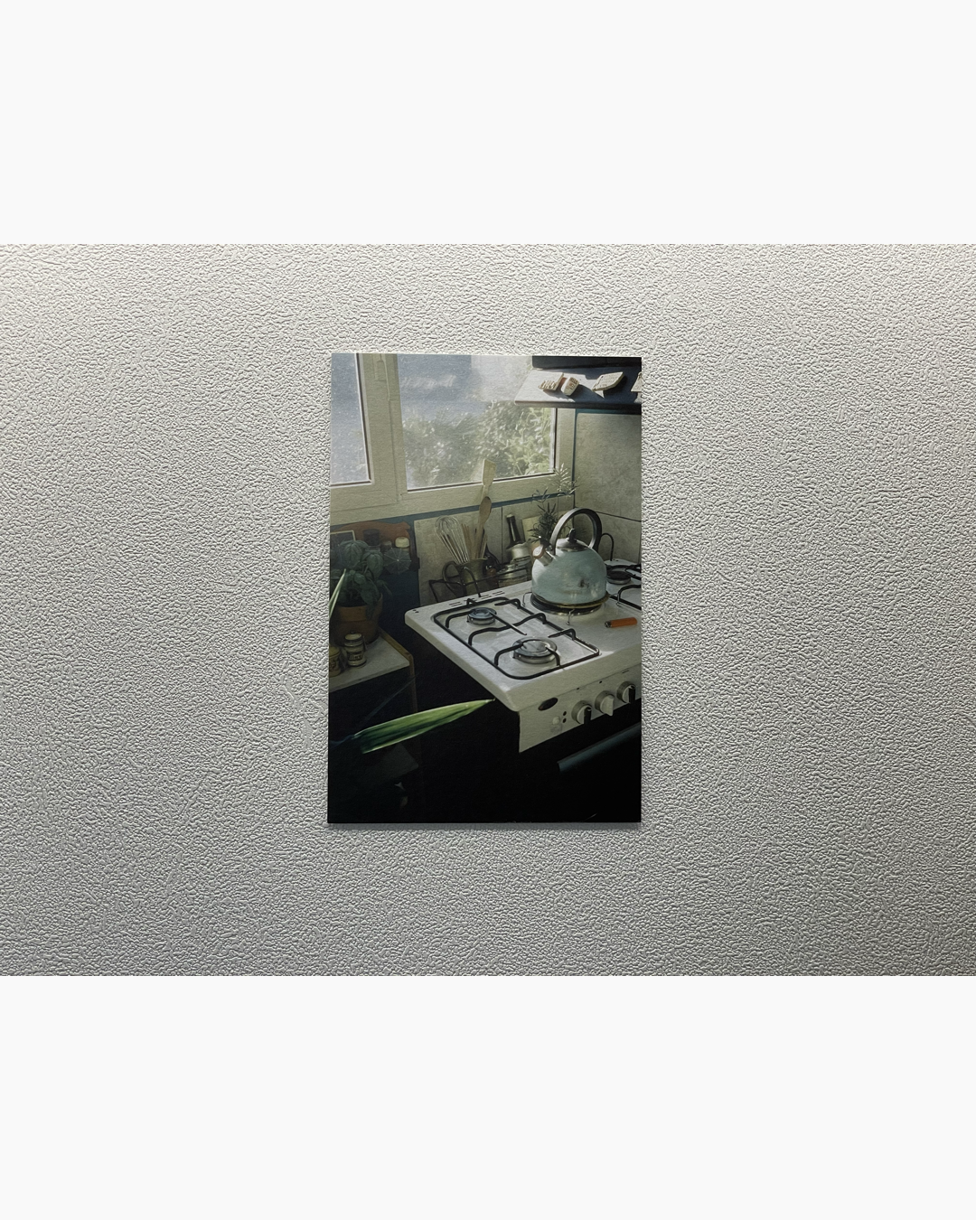 창문 옆 주방 가스레인지 위에 주전자가 놓여 있고 주방 도구와 허브 식물이 주변에 배치된 아늑한 주방 공간.