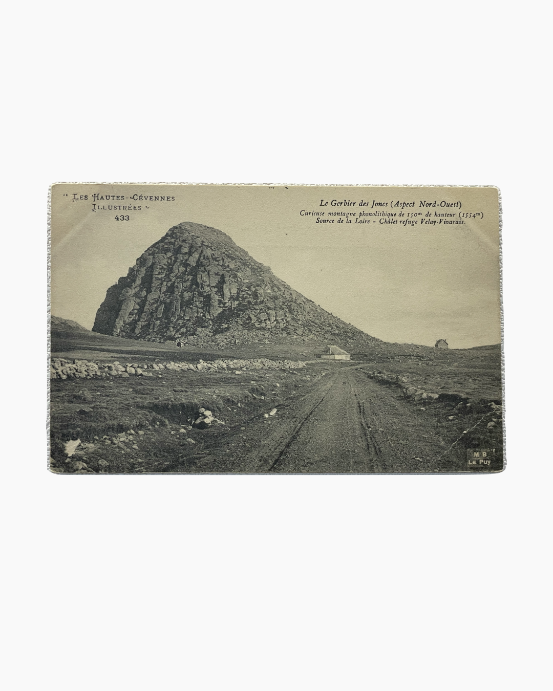 높은 돌무더기 형태의 거대한 바위산이 있습니다. 산 주변에 길과 평평한 초원이 펼쳐져 있으며, 멀리 건물 두 채가 보입니다.