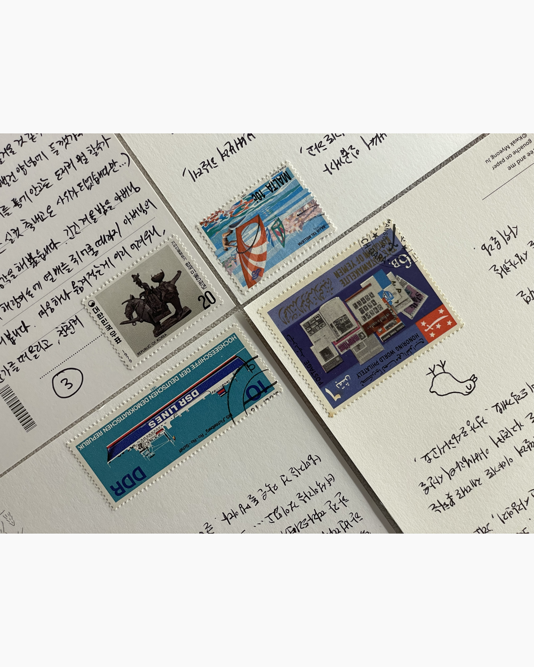 여러 국가의 우표 네 장이 붙은 엽서가 한국어 손글씨 배경 위에 놓여 있음.