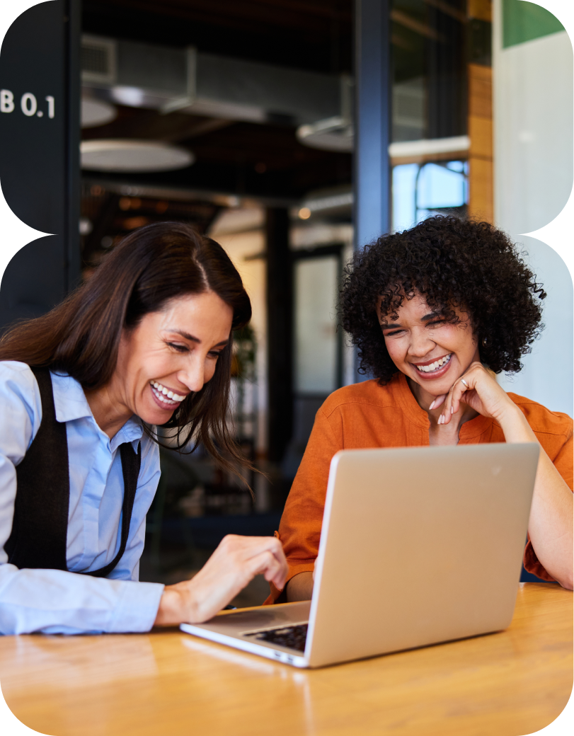 Two women smiling and looking at a laptop screen together at a wooden table in an office setting.