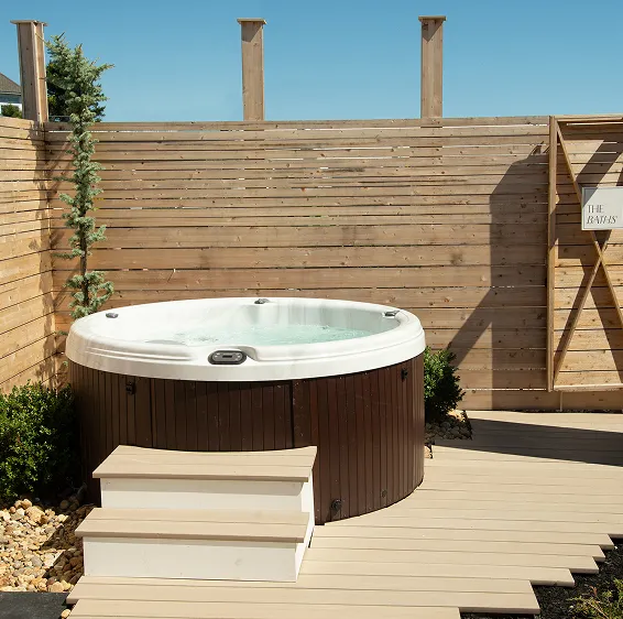 Outdoor hot tub with bubbling water surrounded by wooden decking and a privacy fence under a clear blue sky.