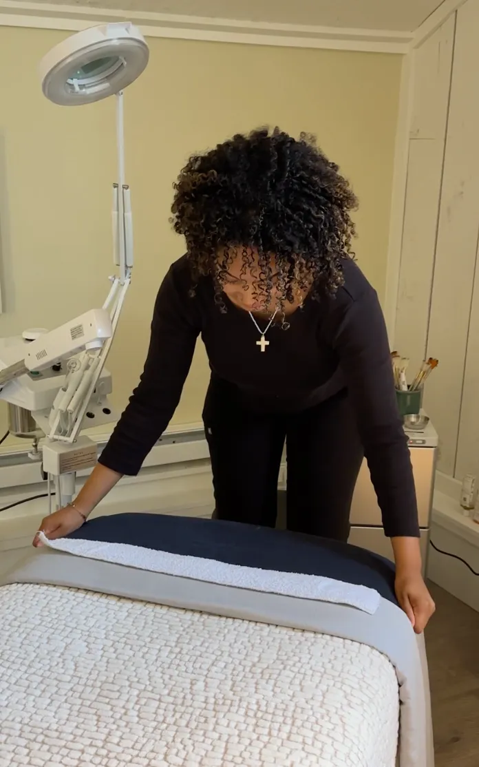 Woman with curly hair and a cross necklace preparing a spa or massage bed with blankets and a towel in a treatment room.