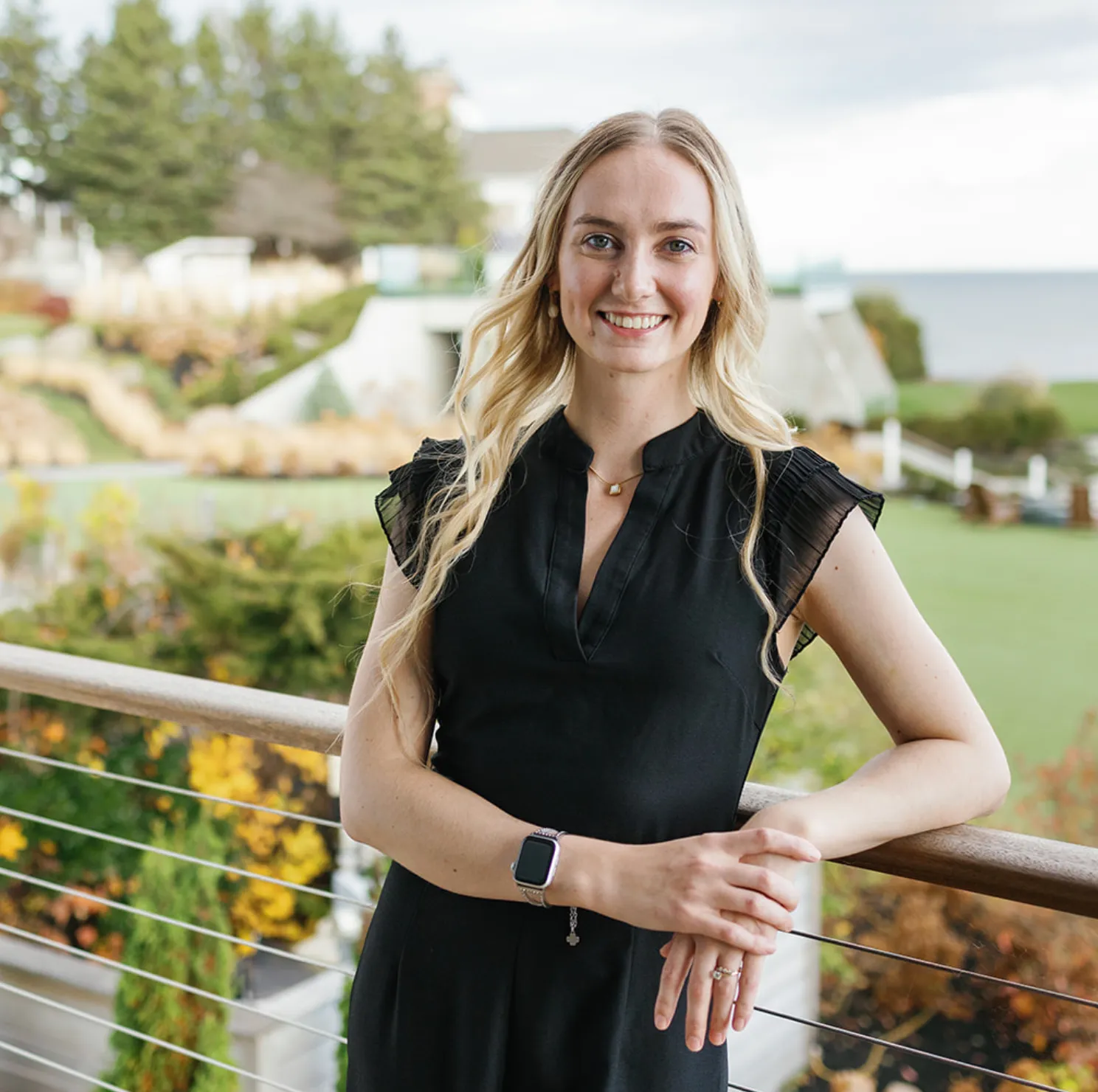 Smiling woman with long blonde hair wearing a black dress and smartwatch, standing outdoors by a railing with greenery and water in the background.