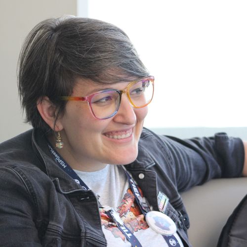 A woman with brown hair and rainbow glasses smiles and looks off to her left