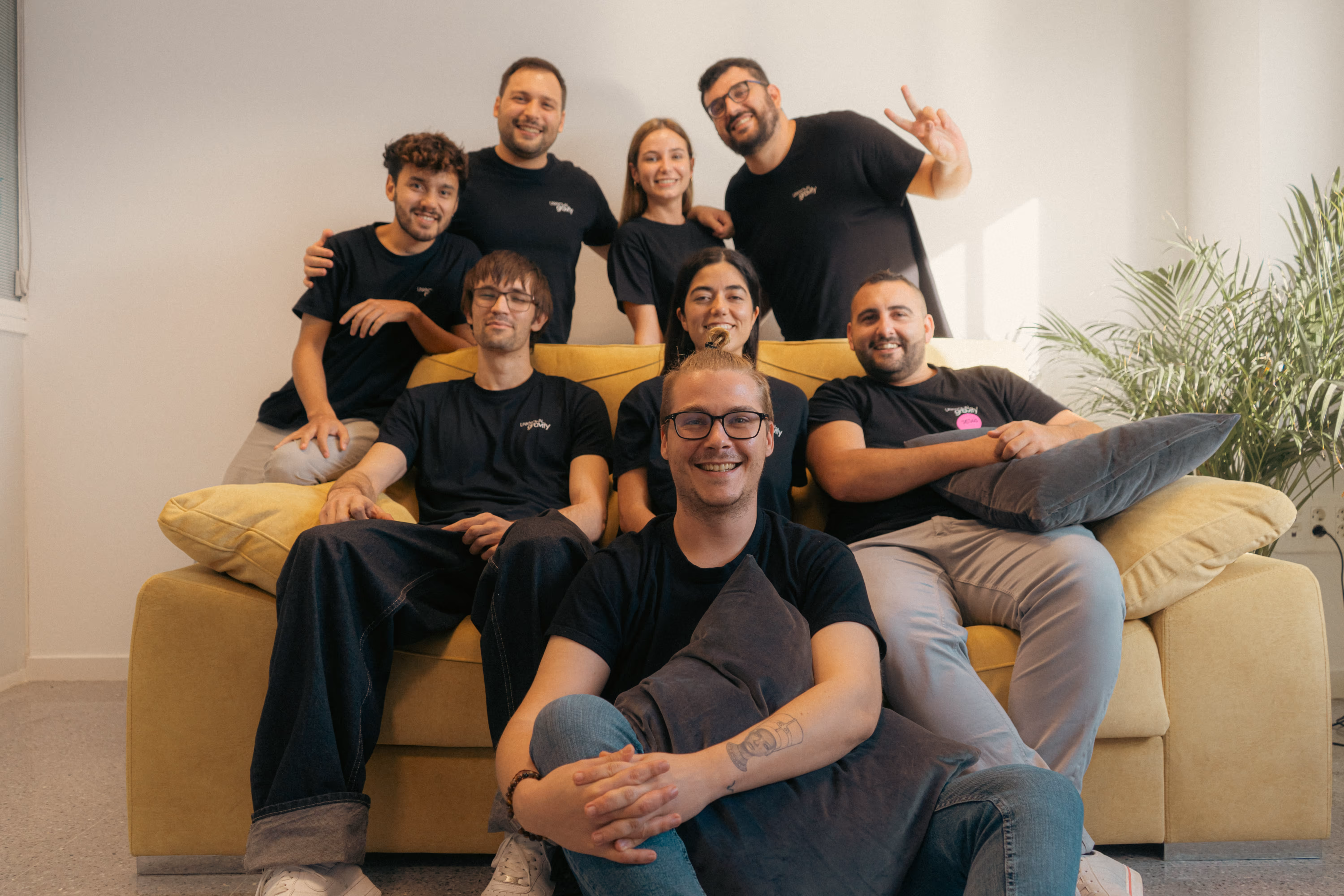 Ocho personas jóvenes sonrientes, cinco sentadas y tres de pie detrás en un sofá amarillo en una sala luminosa con plantas.