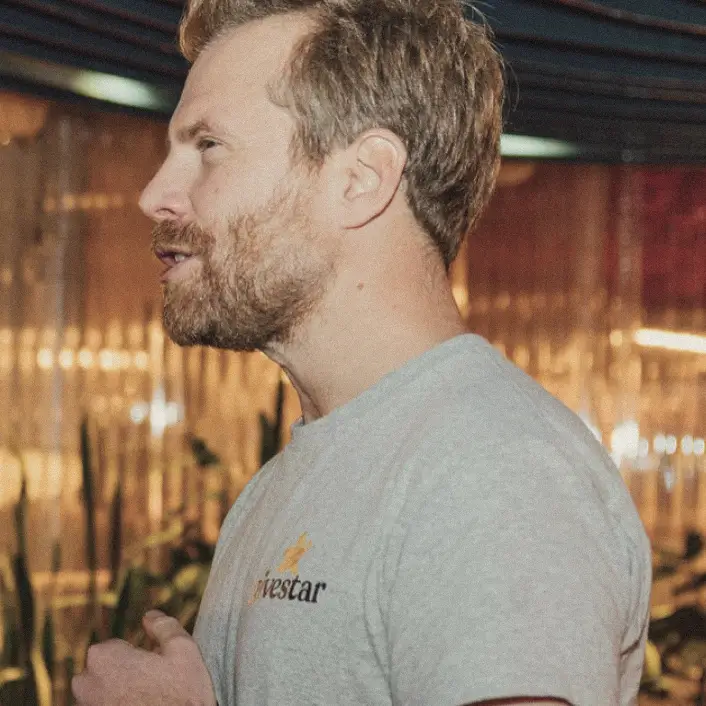 Side profile of a bearded man wearing a light gray t-shirt with a small logo, speaking indoors with blurred background lights.
