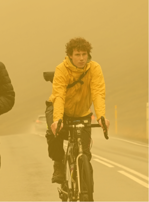 Cyclist wearing a yellow jacket and black pants riding on a foggy road with a wet surface.