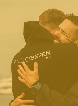 Two men embracing warmly, one wearing a black jacket with the text 'PROJECT SE7EN Huel' on the back.