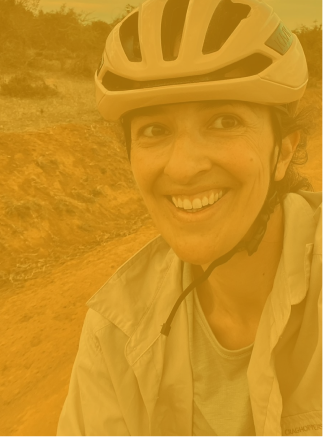 Smiling woman wearing a beige bike helmet and light jacket outdoors on a dirt trail.