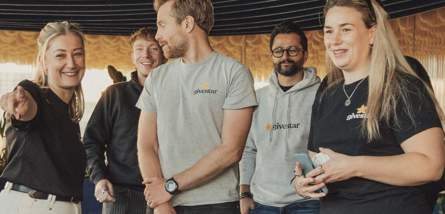 Five smiling people in casual clothing, some wearing shirts with a gold givestar logo, standing indoors.