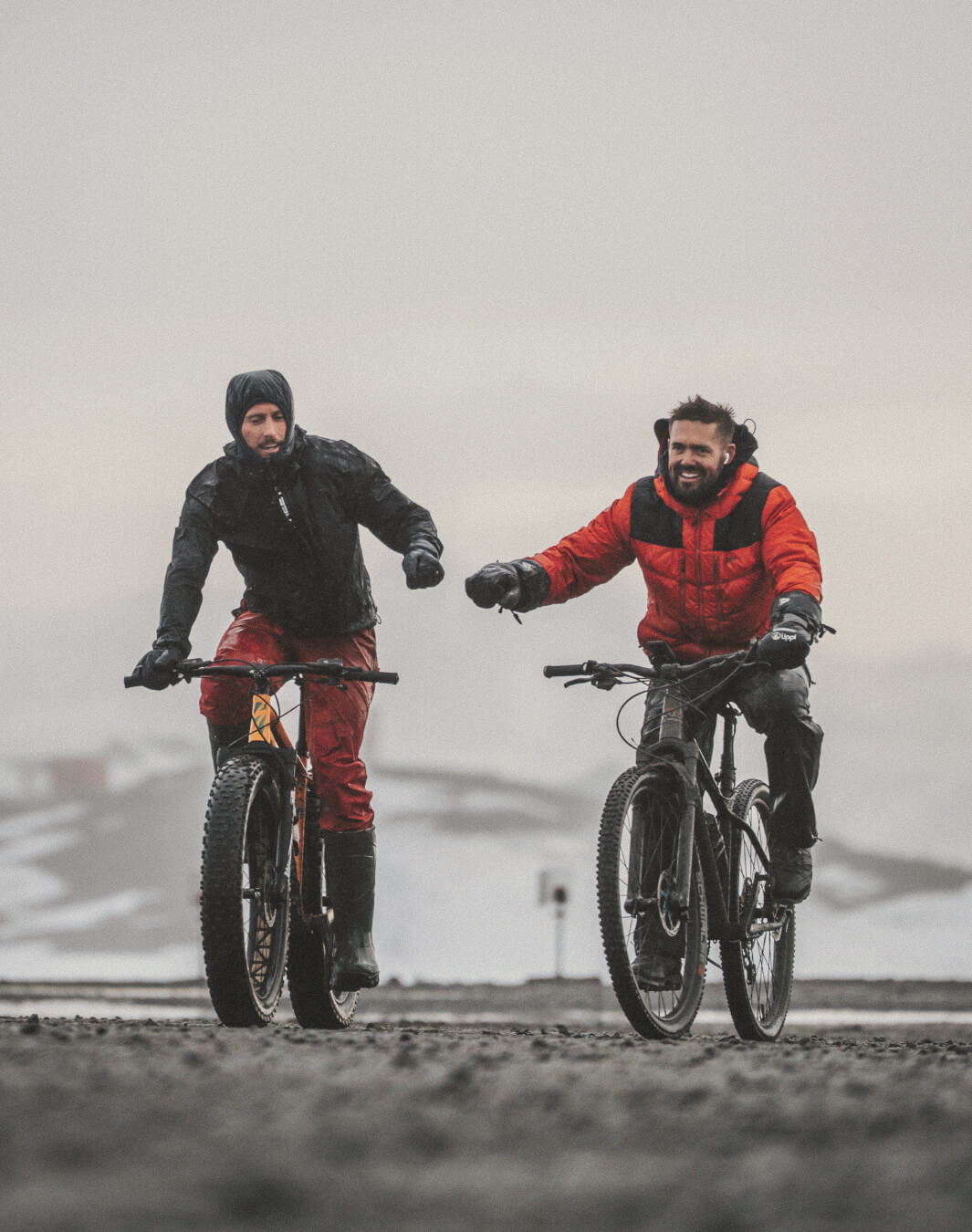 Two men in winter gear fist bump while riding fat tire bicycles on a snowy path.