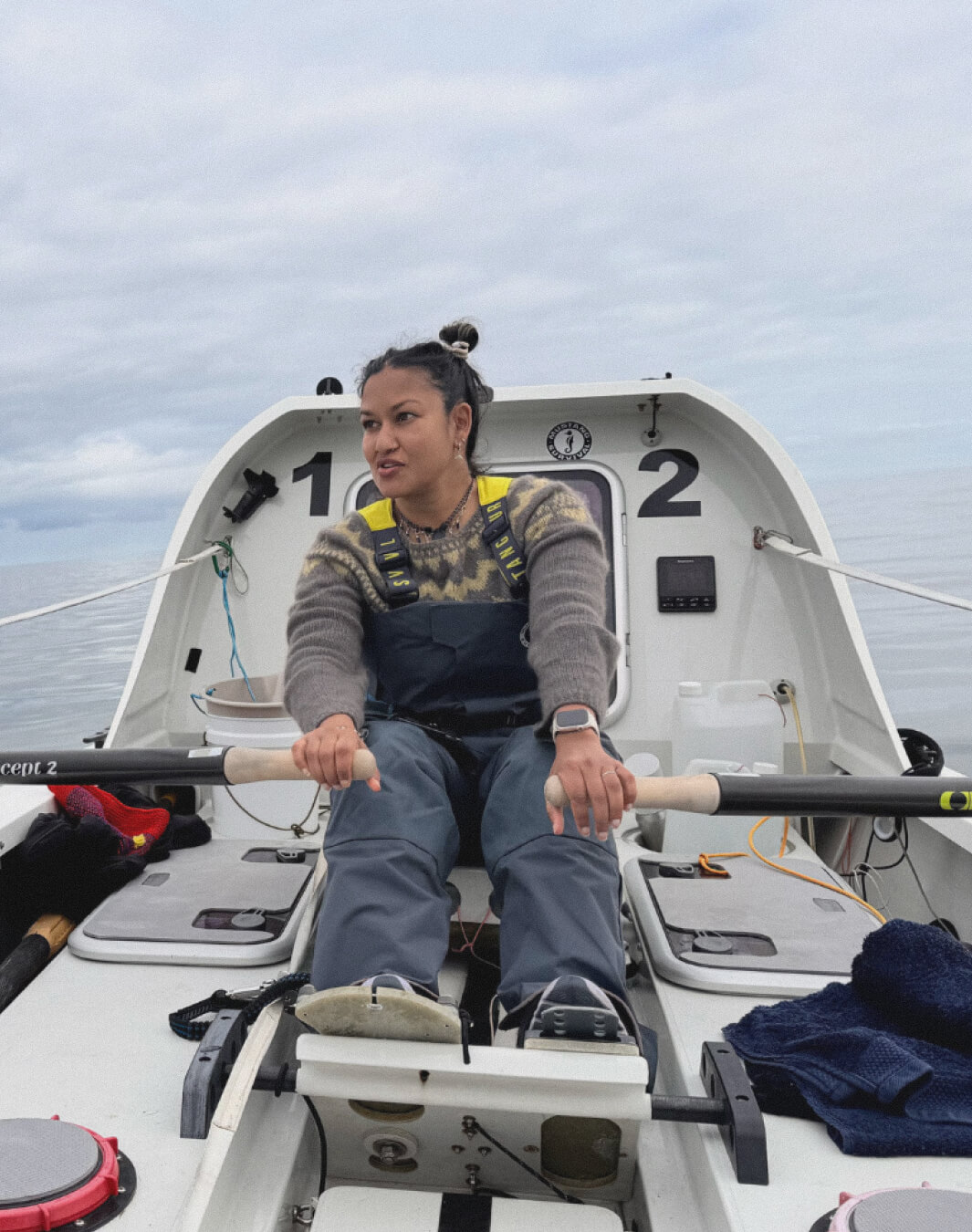 Person rowing a small boat on calm water wearing gray sweater and waterproof pants.