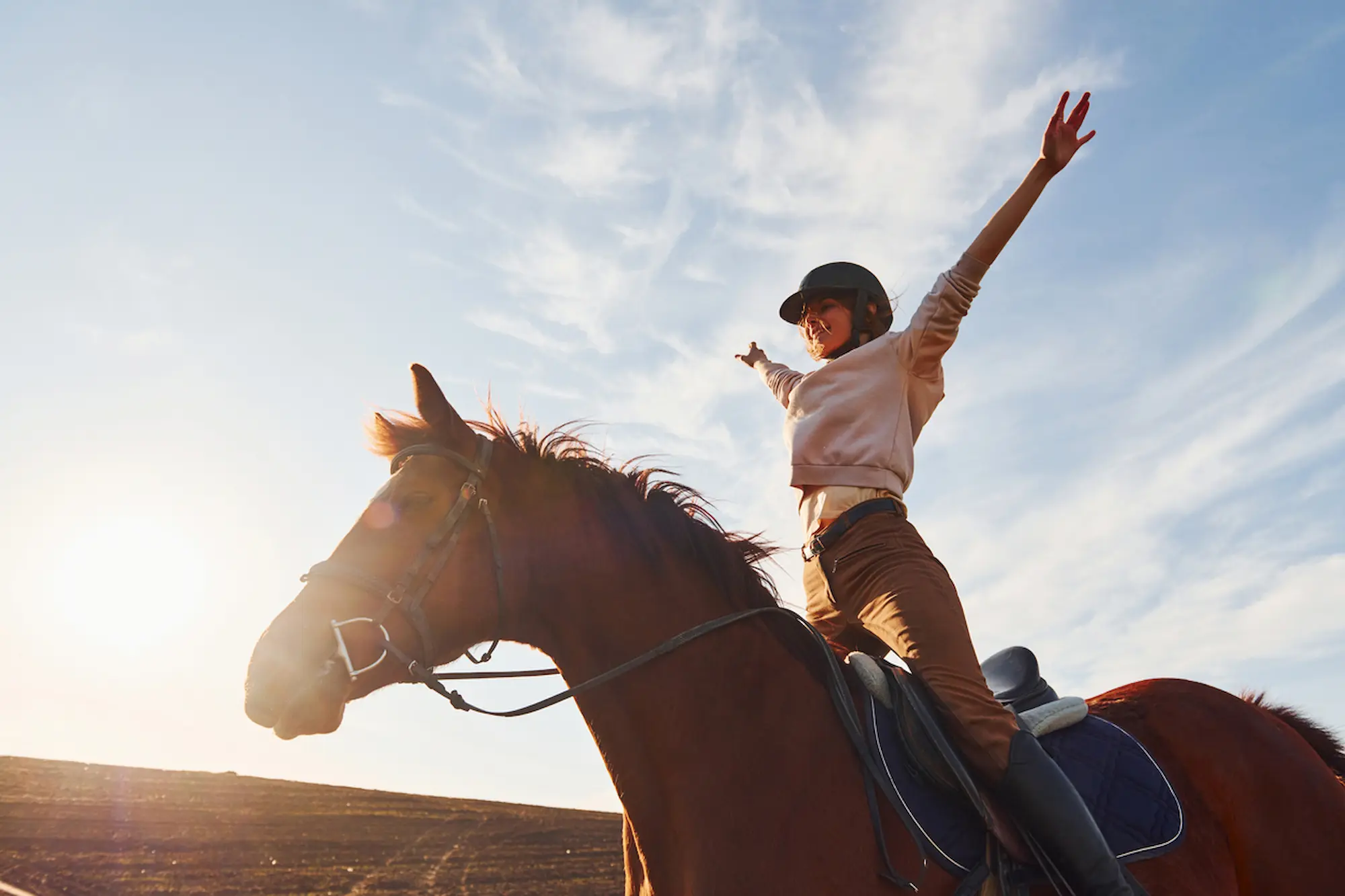 Girl riding a brown horse during sunset with her arms in the air