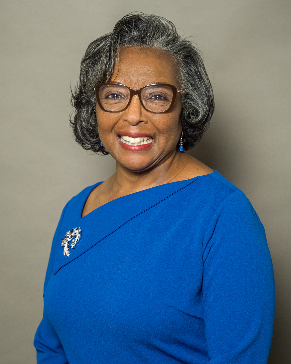 Smiling older woman with gray hair and glasses wearing a blue dress with a decorative brooch.