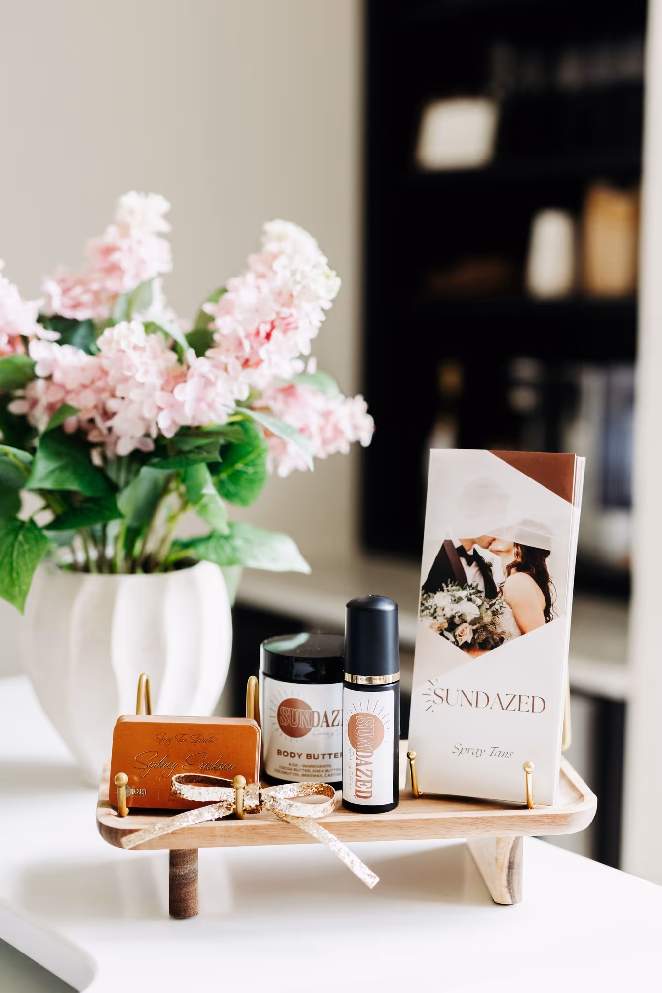 Wooden tray on table displaying Sundazed spray tan products with a vase of pink flowers in the background.
