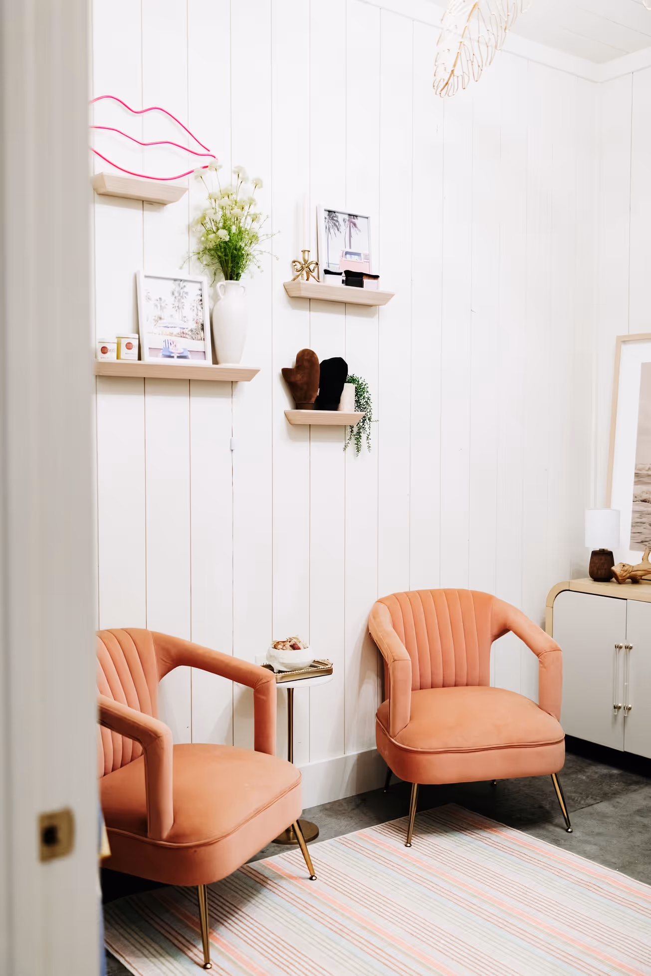 Cozy corner with two peach velvet armchairs, a striped rug, floating shelves with decor, and a white paneled wall.