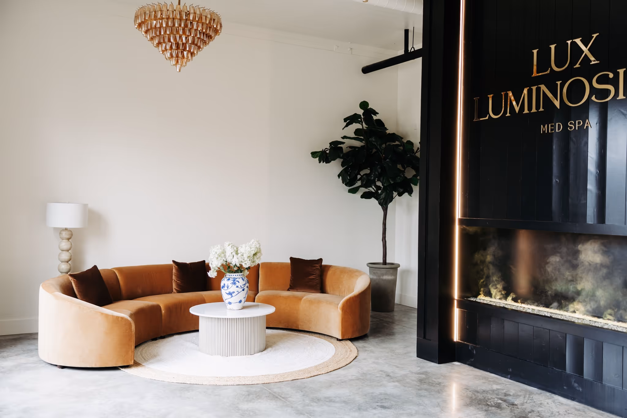 Elegant spa lounge with a curved beige sofa, round white coffee table with floral vase, floor lamp, large potted plant, and modern fireplace.