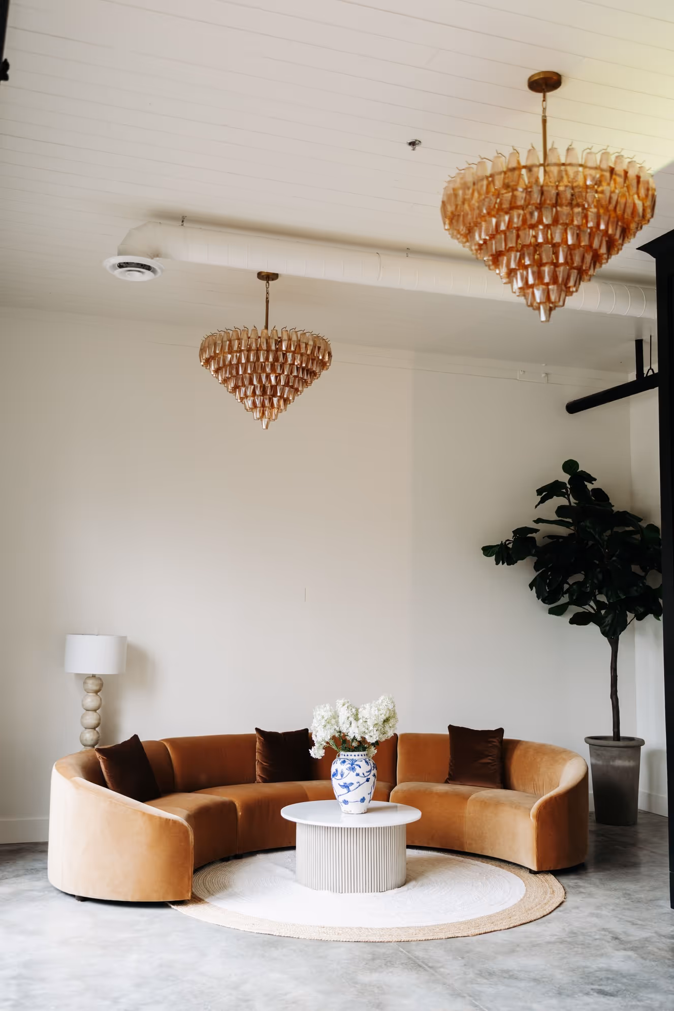Curved tan velvet sofa with dark brown pillows around a round white table holding a blue and white floral vase with white flowers, under two amber glass chandeliers in a modern room with a floor lamp and potted plant.