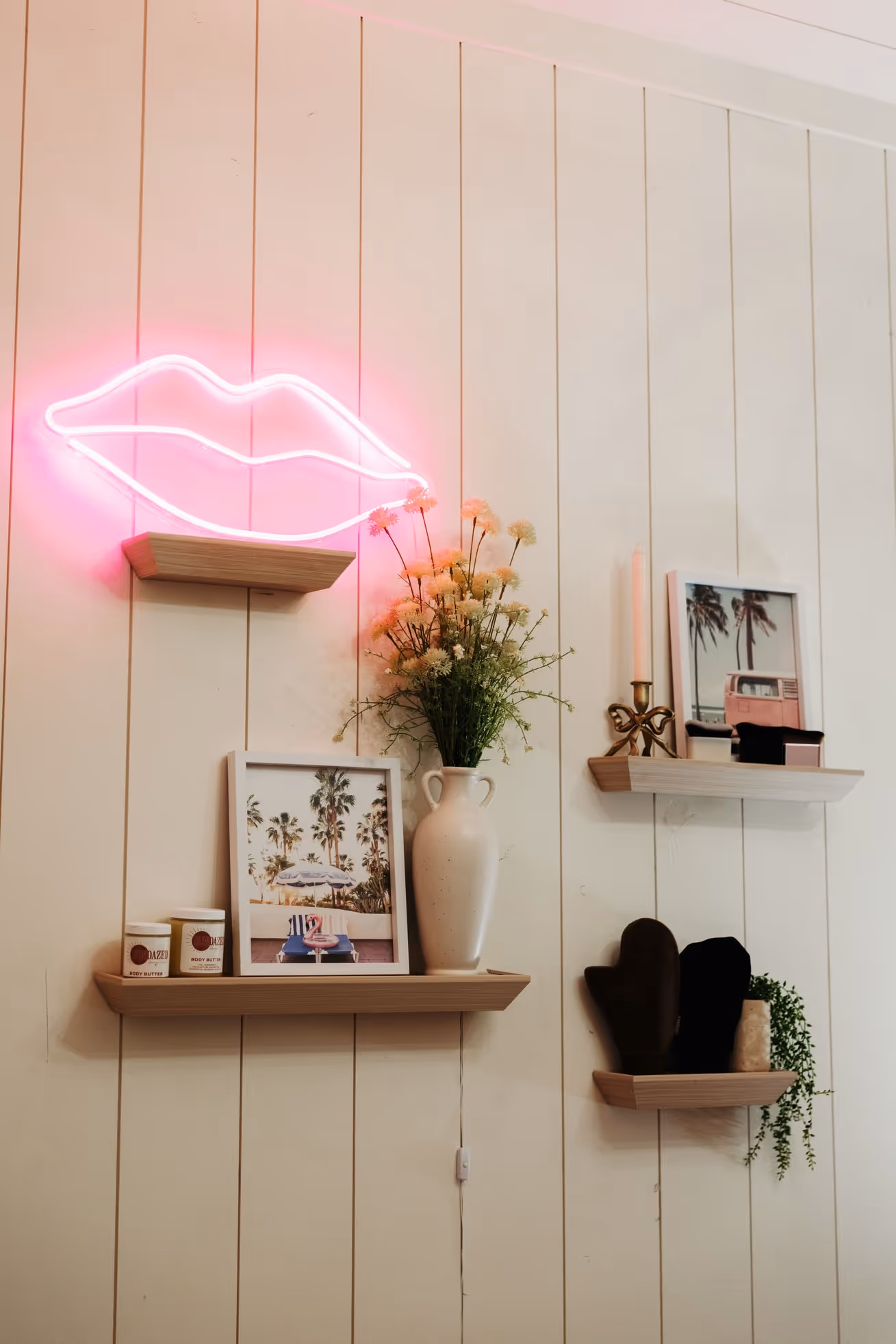 Wall with three wooden shelves holding framed photos, a vase with flowers, body butter jars, candles, mittens, and a neon pink lips light.
