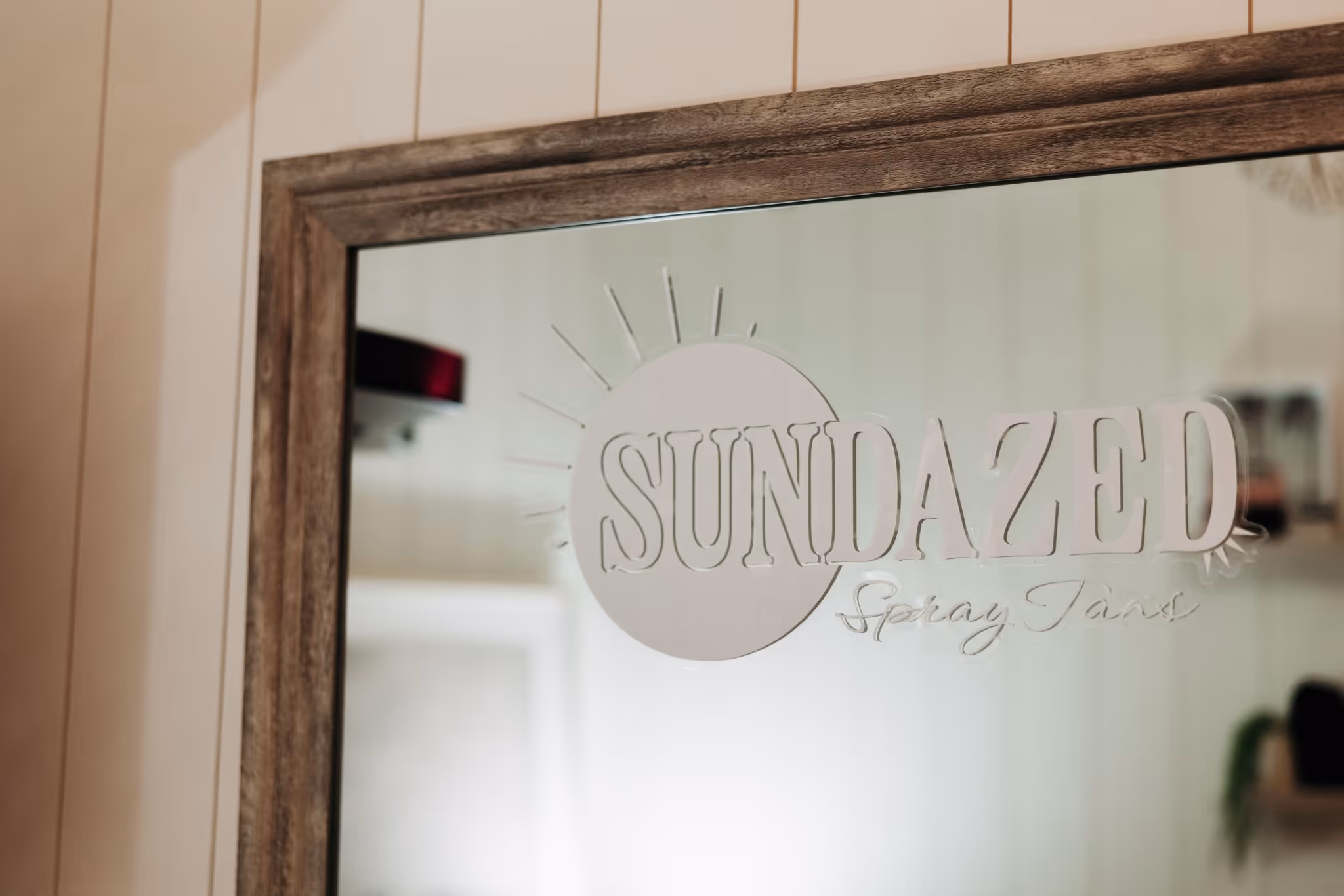Mirror with a wooden frame featuring the logo 'SUNDAZED Spray Tans' and a sun graphic.