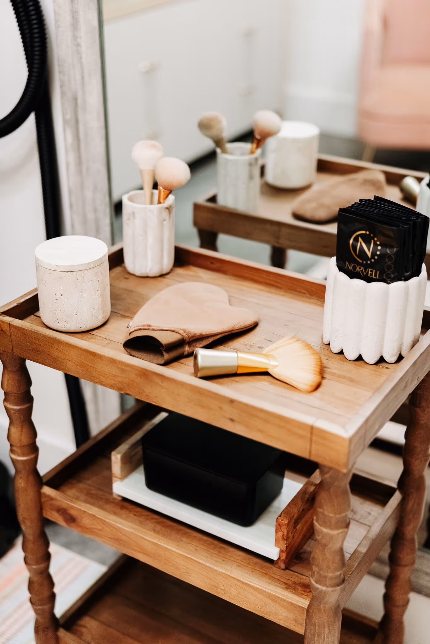 Wooden side table with makeup brushes, beauty mitt, and Norvell tanning wipes in a white ceramic holder.