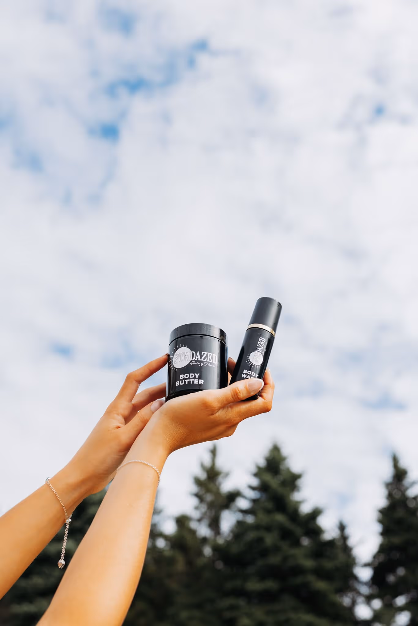 Hands holding black containers of SUNDAZED Body Butter and Body Wash against a partly cloudy sky with pine trees in the background.