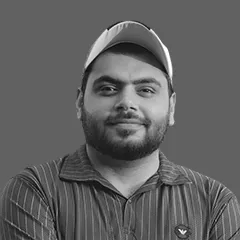 Portrait of a man with a beard and mustache wearing a striped button-up shirt and a baseball cap against a plain background.