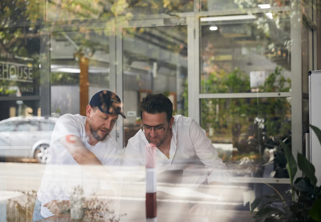 Two men inside a glass-walled office intensely discussing and looking at something on a table.