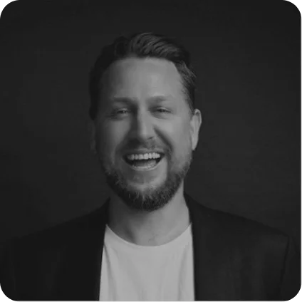 Portrait of a smiling man with a beard and short hair wearing a dark blazer and white shirt against a dark background.