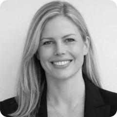 Smiling woman with straight blonde hair wearing a dark blazer against a plain background.