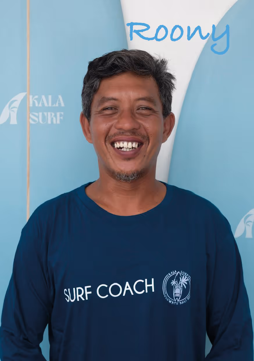 Smiling man with short dark hair wearing a navy blue shirt that says 'Surf Coach' standing in front of turquoise surfboards.