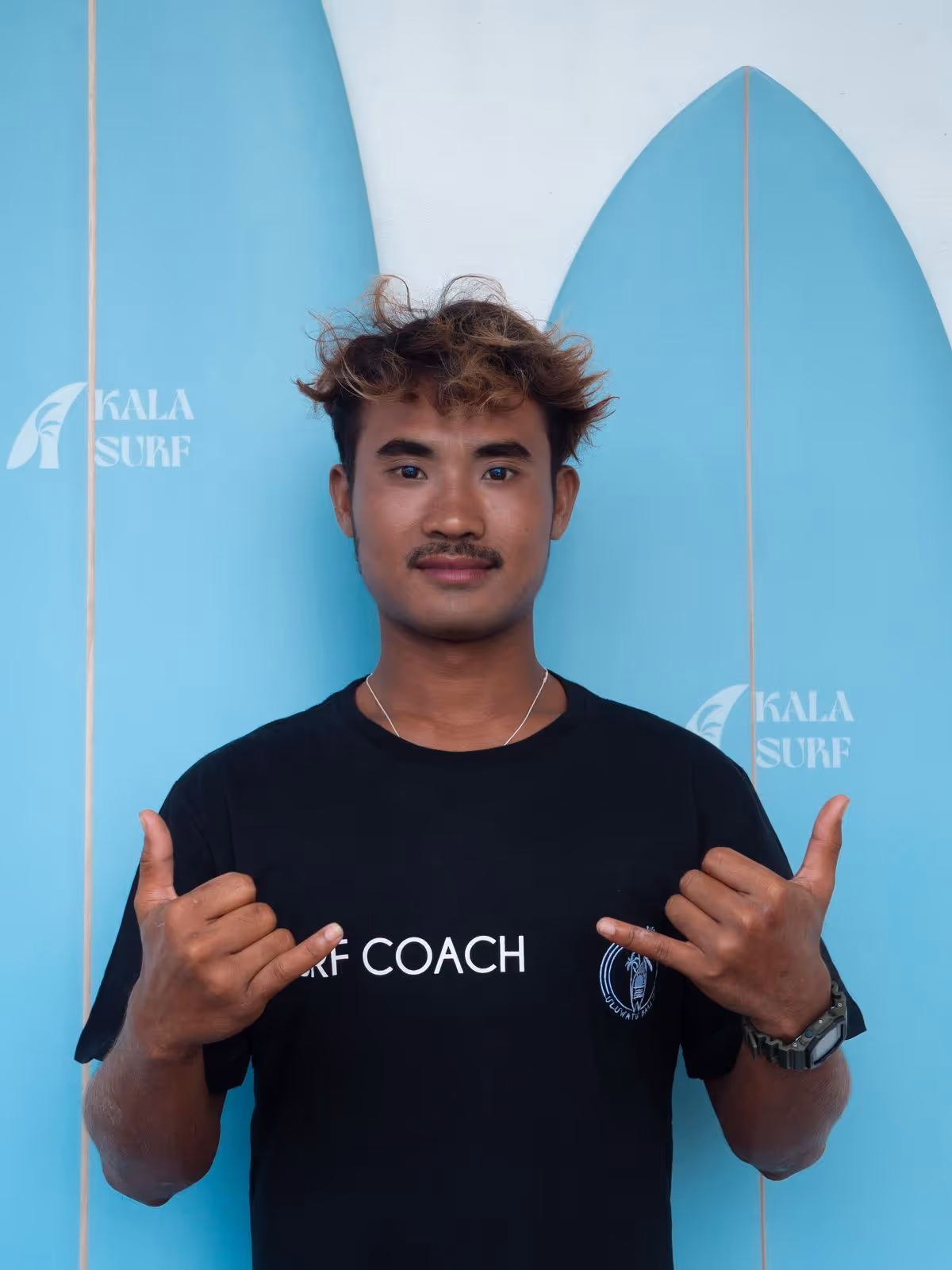 Young man in a black 'Surf Coach' t-shirt making shaka signs with both hands, standing in front of two blue surfboards.