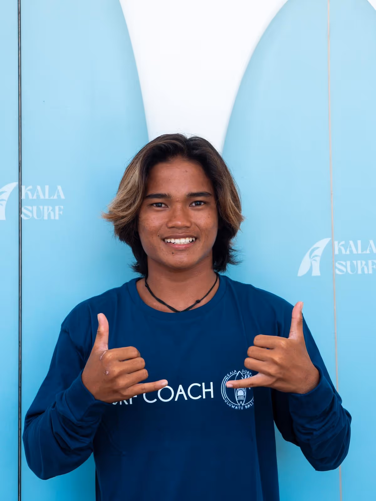 Smiling young man with shoulder-length hair wearing a blue long-sleeve shirt reading 'SURF COACH' and making shaka hand signs in front of a blue and white surfboard.