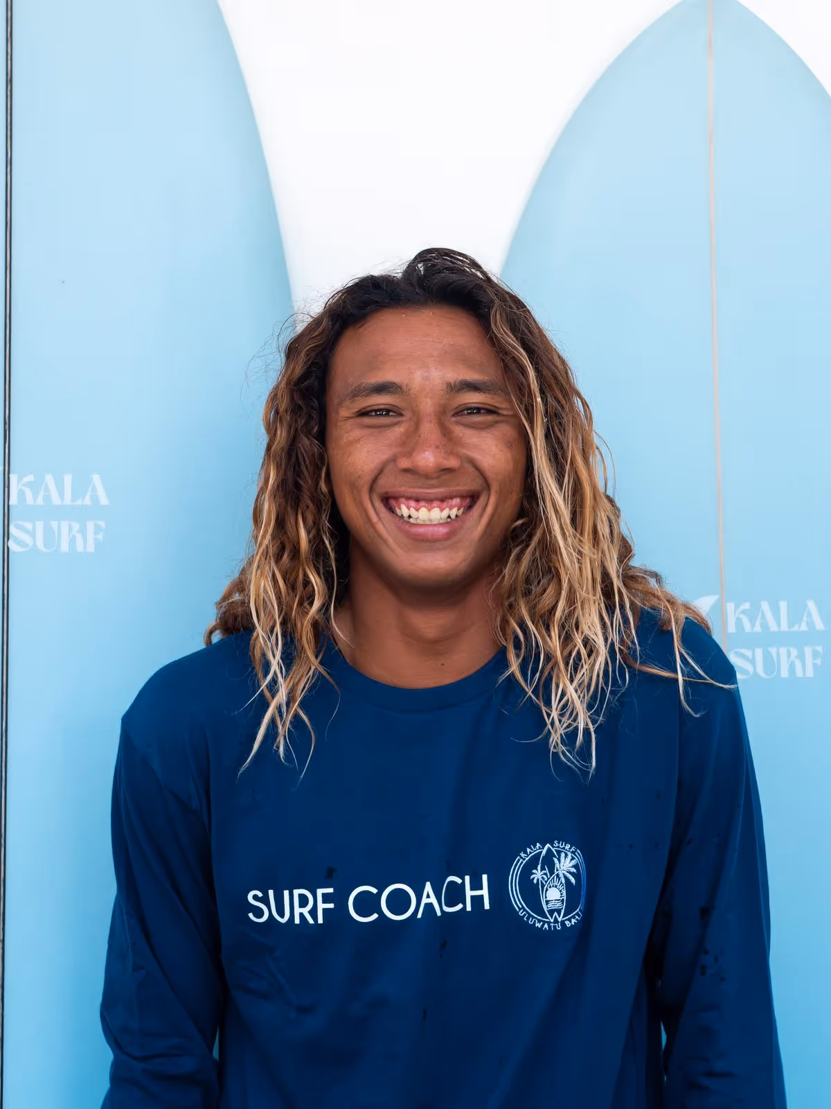 Smiling surf coach with long curly hair wearing a navy blue shirt in front of blue surfboards.