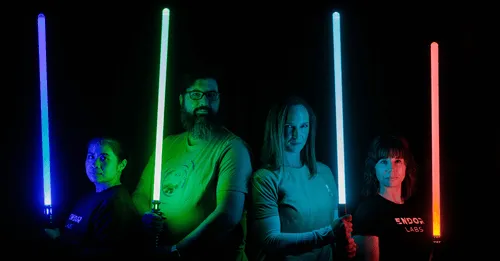 Four people standing in front of a black background, each holding a glowing lightsaber in blue, green, cyan, and red colors.