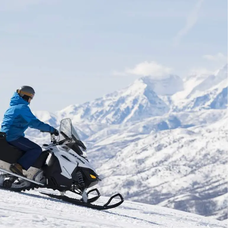 A person riding a snowmobile up a snowy mountain slope, surrounded by vast snow-covered peaks under a clear sky.