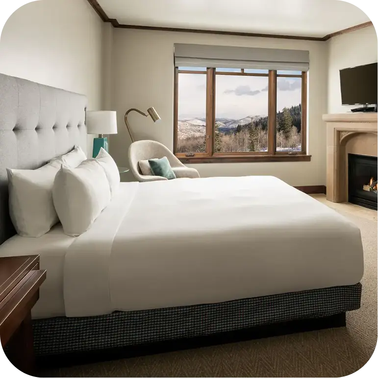 A cozy hotel room featuring a large bed, armchair, fireplace, and a window with a view of snow-covered trees and mountains.