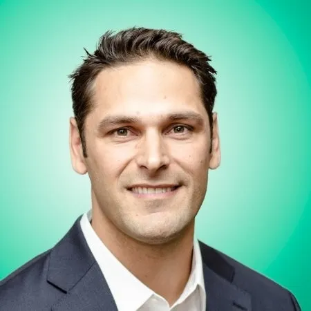 Portrait of a smiling man with dark hair wearing a white shirt and dark blazer against a teal gradient background.