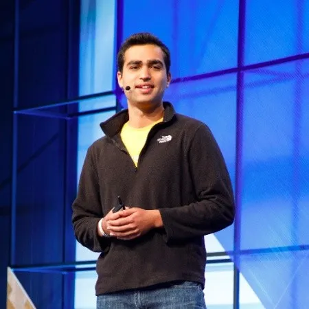 Man wearing a black fleece jacket and headset microphone speaking on a stage with blue background panels.