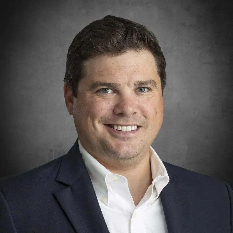 Smiling man with short dark hair wearing a navy blazer and white shirt against a dark gray background.